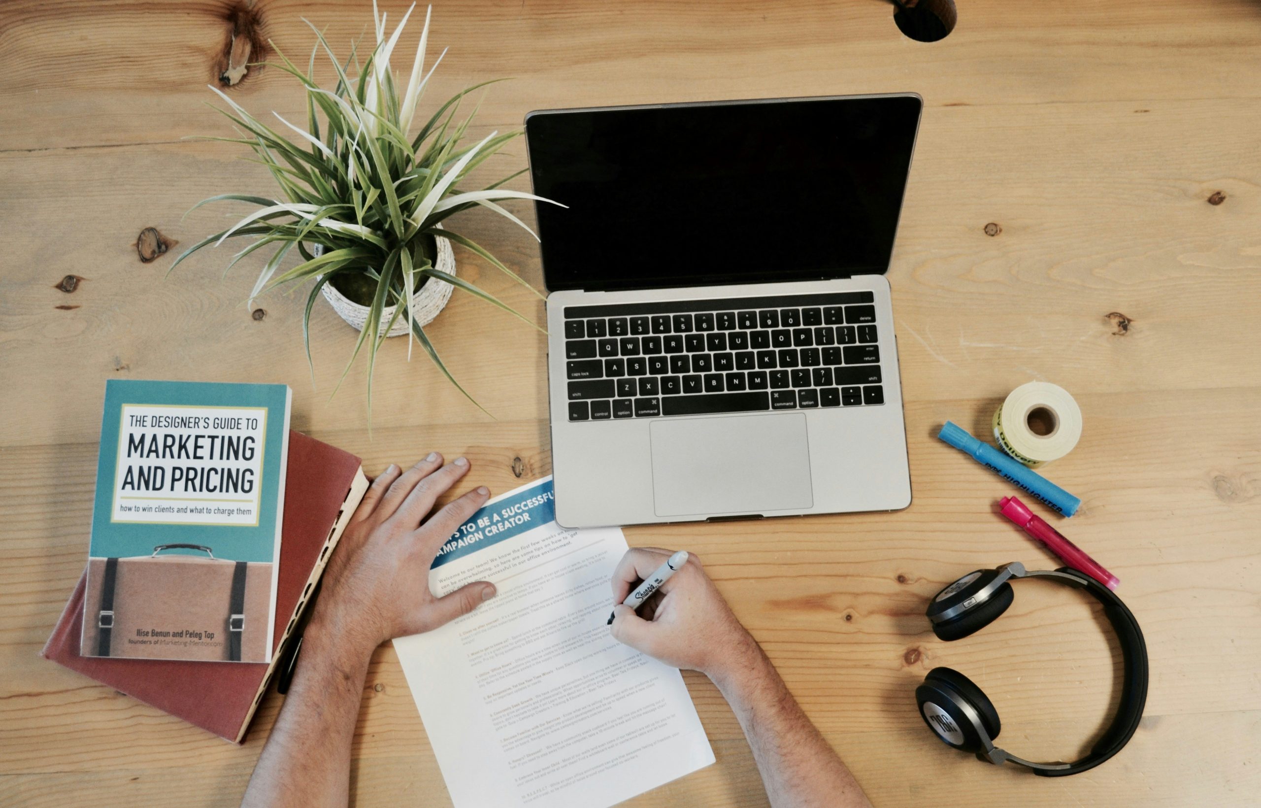 Ein Schreibtisch mit Laptop, Kopfhörern, Pflanze, Zetteln und einem Buch mit der Aufschrift Marketing