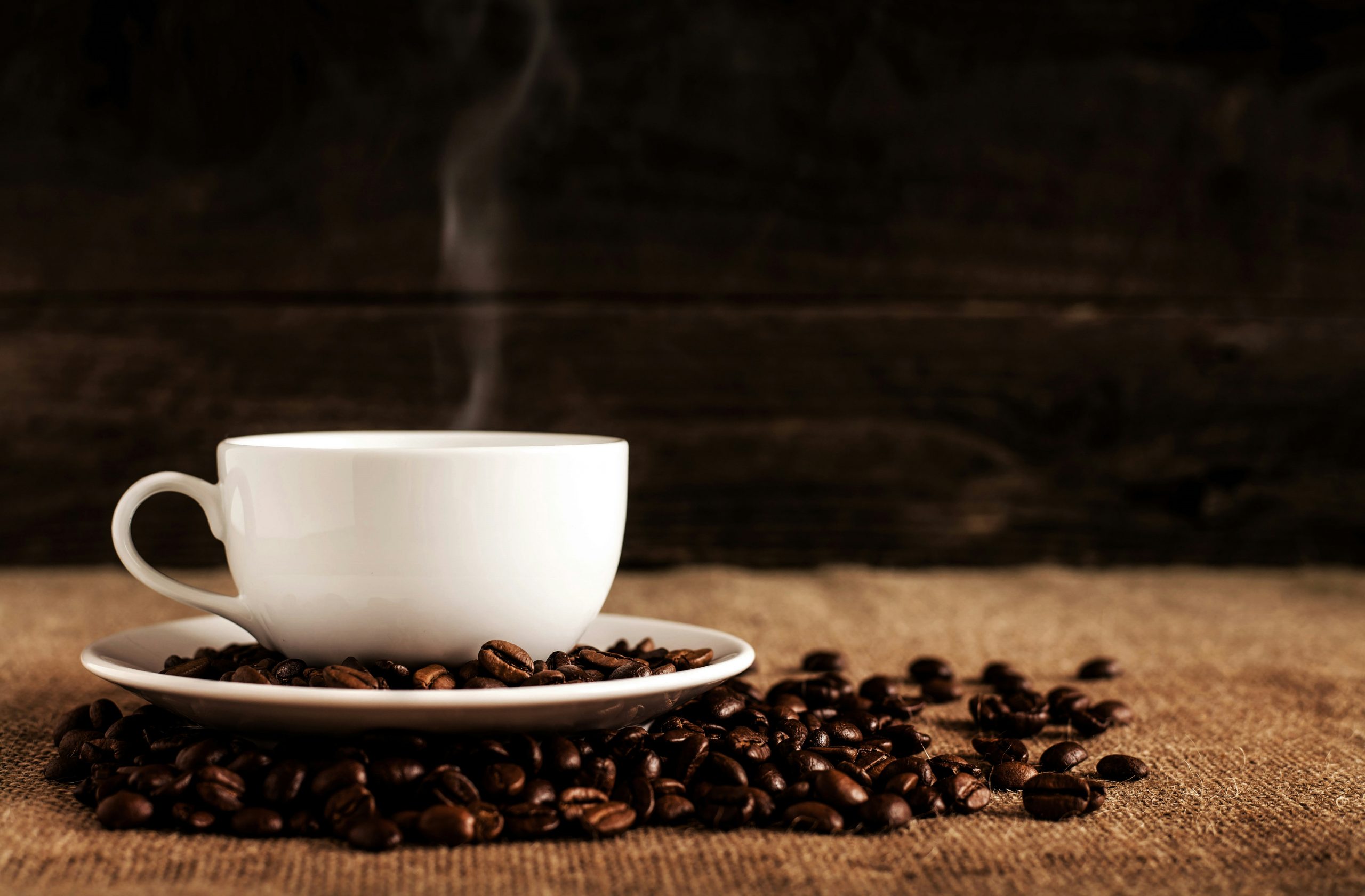 Ein Kaffeebecher mit dampfendem Inhalt und diversen Kaffeebohnen daneben