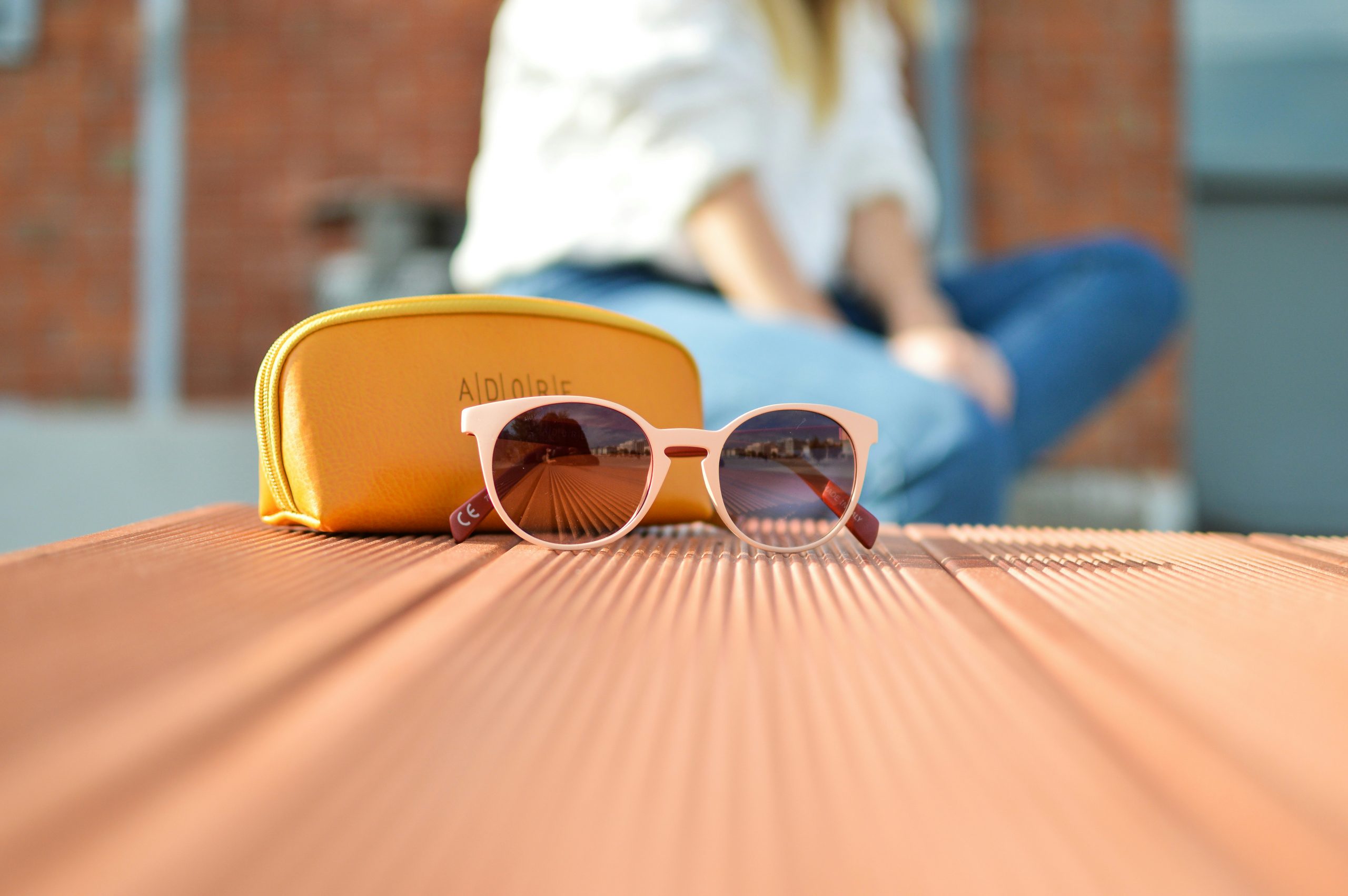 Eine Sonnenbrille und Etui auf einem Holztisch, im Hintergrund sitzt eine Frau in weißer Bluse und Jeans